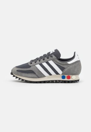 Zapatilla Adidas gris con rayas blancas, malla texturizada, paneles de ante y suela estriada, con detalles circulares en azul, rojo y blanco en el talón.