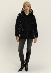 Zwarte glanzende pufferjas met diamanten quiltpatroon, een capuchon en een rits aan de voorkant. Gecombineerd met donkere jeans en hoge zwarte stiletto's tot de knie.