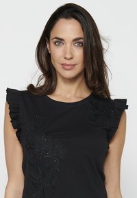 T-shirt noir avec des manches volantées et une broderie florale agrémentée de perles sur le devant, fabriqué en tissu doux et respirant.