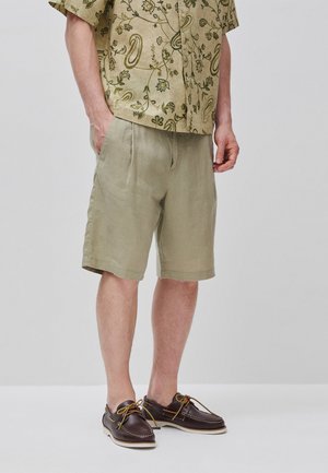Hombre con camisa de manga corta beige con estampado floral, pantalones cortos caqui claros y zapatos náuticos de cuero marrón con cordones amarillos.