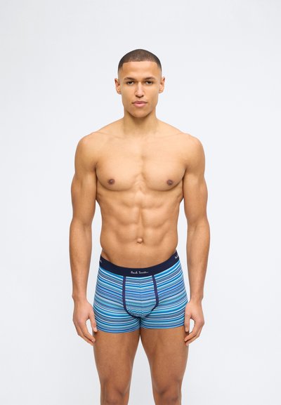 Homme musclé debout, face à l'avant, portant un boxer rayé bleu avec une ceinture bleue marine, sur un fond clair uni.