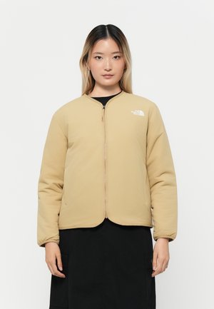 Beige isolert jakke med rund hals, frontglidelås og en liten hvit logo på venstre bryst. Glatt tekstur og polstret design.