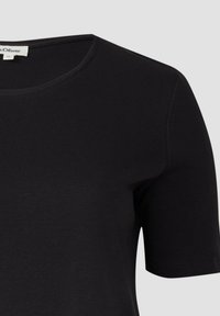 Schwarzes Kurzarm-T-Shirt mit Rundhalsausschnitt und einem kleinen weißen Markenlabel im Kragen auf einem schlichten hellen Hintergrund.