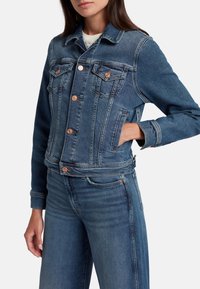 Giacca di jeans blu scuro con un design aderente, dotata di bottoni a pressione, tasche sul petto e cuciture a contrasto. Tessuto liscio con un aspetto leggermente scolorito.