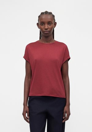 Donna con i capelli intrecciati che indossa una t-shirt a maniche corte rosso scuro e pantaloni blu scuro, in piedi davanti a uno sfondo chiaro e uniforme.