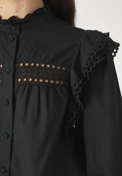 Svart blus med en texturerad panel med cirkulära utskärningar och intrikata spetsdetaljer på axlarna. Knäppning vid halsen.