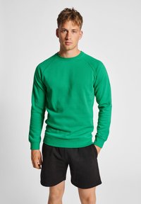 Sweatshirt vert à col rond et manches raglan, fabriqué en un mélange de coton lisse, associé à un short noir.