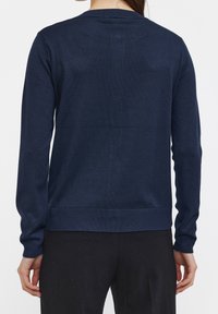 Žena v pohodlném navy modrém svetru s dlouhým rukávem a černých kalhotách, zachycena z pohledu zezadu na bílém pozadí.