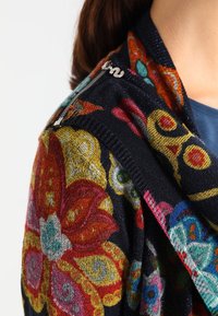 Närbild av en axel med en mörk tygtröja som har stora färgglada blommönster och en silverdragkedja i kragen.