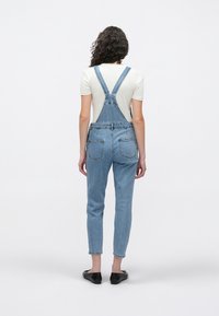 Femme aux cheveux bouclés portant une salopette en denim bleu clair et un t-shirt blanc à manches courtes, debout de dos sur un fond uni.