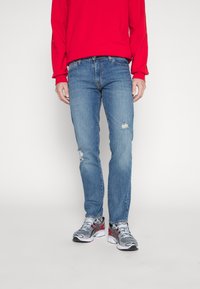 Jean en denim bleu clair avec un léger effet vieilli, coupe ajustée, associés à une chemise rouge à manches longues et des chaussures de sport grises.