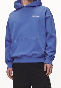 Hombre con una sudadera azul con bolsillo frontal y logo blanco de "PEGADOR", combinada con pantalones cargo negros.
