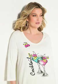 Camisetta bianca con scollo a barca e maniche a 3/4, caratterizzata da una grafica colorata di un cocktail con accenti di frutta e la parola "Cocktail" in testo nero.