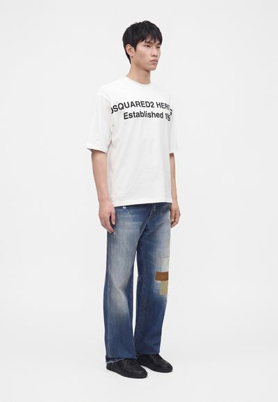 Νέος άνδρας που φοράει λευκό φαρδύ T-shirt με μαύρο κείμενο και ξεβαμμένο μπλε τζιν με μπαλώματα, στέκεται μπροστά σε απλό λευκό φόντο.