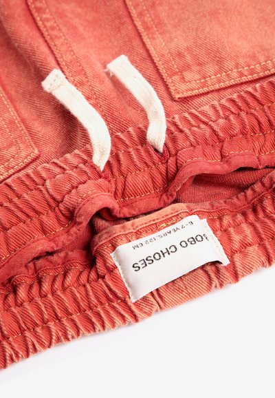 Gros plan sur une ceinture élastique couleur corail avec des cordons blancs et une étiquette en tissu indiquant la taille et la marque.