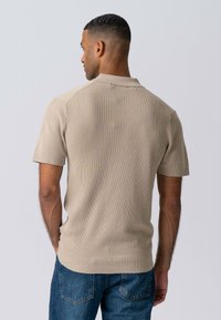 Beige ribbet polo-shirt med korte ærmer og krave, med en tekstureret stofdetalje, parret med blå denimjeans.