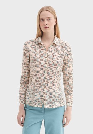 Frau trägt ein cremefarbenes Langarmshirt mit wellenförmigen roten und blauen Linien, kombiniert mit hellblauen Hosen, und steht vor einem einfarbigen Hintergrund.