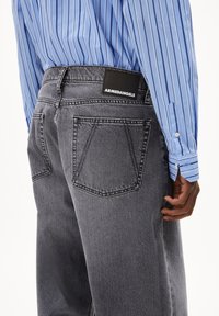 Grijze denim jeans met een relaxte fit, voorzien van achterzakken en een zwart ARMEDANGELS-label op de tailleband. Gestreept blauw shirt en hand zichtbaar.