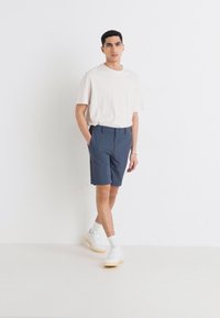 Light-colored, short-sleeve t-shirt paired with dark blue shorts. Model wears white sneakers and crew-length socks, standing against a white background.
