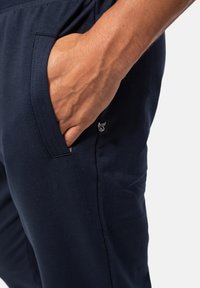 Donkerblauwe sportbroek met een gladde textuur, voorzien van een zijzak en een klein logo-label. Het ontwerp is functioneel en aansluitend.
