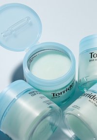 Klare, runde plastikbeholdere med hudplejeprodukt med lyseblå låg. En beholder er åben, hvilket afslører et gel-lignende, mintgrønt stof indeni.