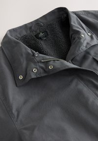 Veste grise en tissu avec un col doublé de fourrure noire douce, boutons-pression et fermeture éclair à l'avant. Présente une texture lisse et un design décontracté.