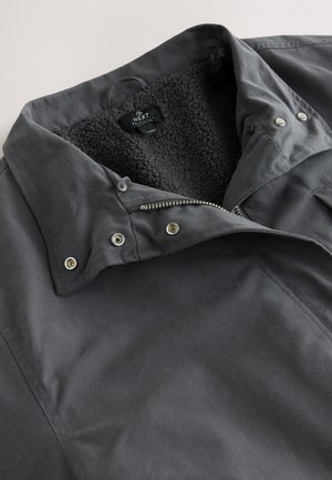 Veste grise en tissu avec un col doublé de fourrure noire douce, boutons-pression et fermeture éclair à l'avant. Présente une texture lisse et un design décontracté.