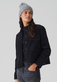 Zwarte corduroy jas met twee voortassen, knoopsluiting en zachte textuur, gecombineerd met een grijze gebreide beanie.