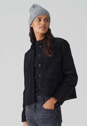 Zwarte corduroy jas met twee voortassen, knoopsluiting en zachte textuur, gecombineerd met een grijze gebreide beanie.