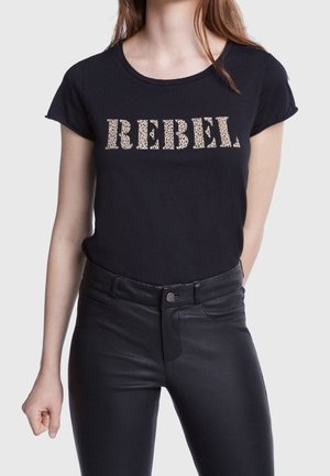 Kobieta w czarnej koszulce z napisem "REBEL" w panterkę i czarnych, skórzanych, wysokich spodniach, stojąca na jednolitym tle.