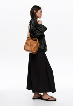 Mujer con chaqueta de cuero negra y falda larga negra, parada y usando chanclas negras, llevando un bolso de cuero marrón con correa beige al hombro.