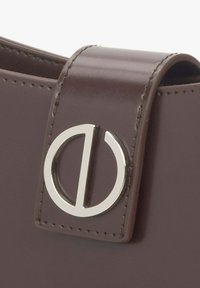 Detalj smeđe kožne torbe s okruglim srebrnim akcentom na hardveru i zatvaranjem na preklop, ističući elegantan dizajn i majstorsku izradu.