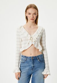 Weißes kurzes Oberteil mit Rüschensaum am Hals und langen Ärmeln, mit einem gebundenen Frontverschluss und einem strukturierten karierten Muster. Kombiniert mit blauen Jeans.