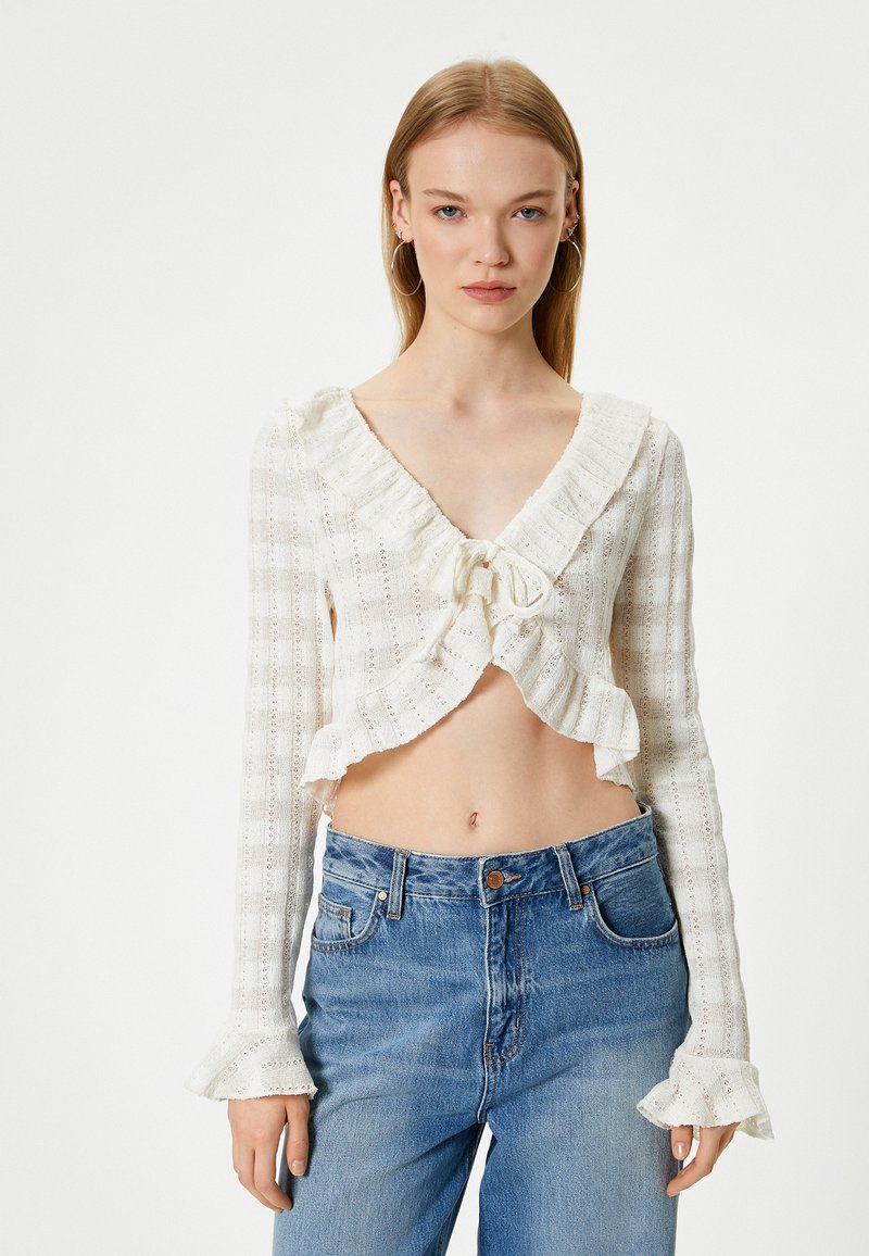 Hvid cropped top med flæser ved halsen og lange ærmer, med en knyt-front og et struktureret ternet mønster. Sammen med blå jeans.