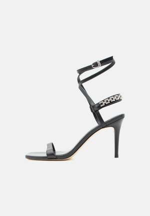 Sandale noire à talon haut avec fine bride à la cheville, bride médiane décorée de maillons de chaîne argentés, et design à bout ouvert.