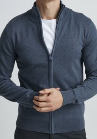 Mężczyzna w granatowym swetrze zapinanym na zamek, noszącym pod nim białą koszulkę, z rękami skrzyżowanymi na brzuchu, na tle jednolitego jasnego tła.