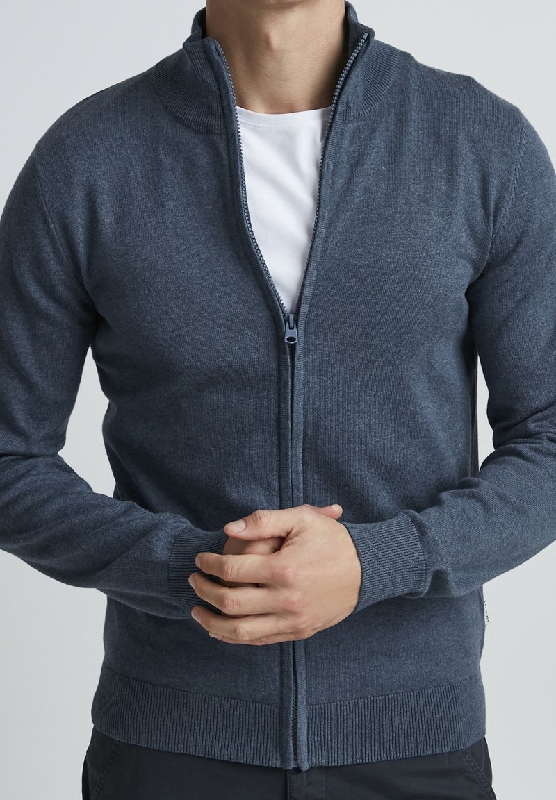 Mężczyzna w granatowym swetrze zapinanym na zamek, noszącym pod nim białą koszulkę, z rękami skrzyżowanymi na brzuchu, na tle jednolitego jasnego tła.