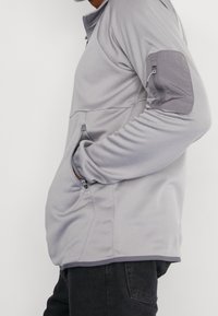 Svetle šedý pulovr s dlouhým rukávem a kapsou na zip na rukávu, žebrovaná textura a přímý střih. Obsahuje kontrastní záplaty na loktech.