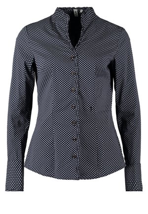 Frau trägt ein schwarz-weiß gepunktetes Hemd mit Knöpfen, weiße Skinny-Hosen und schwarze Stiefeletten, steht mit einer Hand in der Tasche.