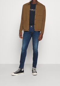 Giacca marrone a bottoni sopra una maglietta blu navy, abbinata a jeans slim blu e sneakers alte nere. Design semplice, tessuti con texture.