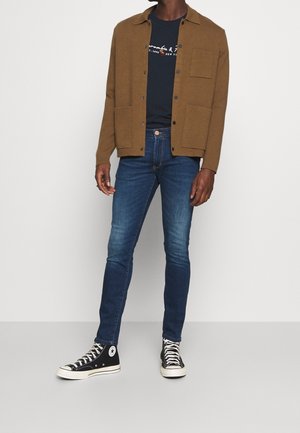 Brązowa kurtka zapinana na guziki na granatowej koszulce, w zestawieniu z wąskimi niebieskimi dżinsami i czarnymi trampkami wysokimi. Prosty fason, teksturowane materiały.