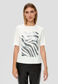 T-shirt blanc à manches courtes avec un motif ondulé en paillettes argentées et noires. Le tissu est lisse ; la coupe est décontractée et légèrement carrée.