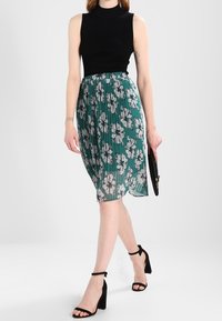 Top senza maniche a coste nere abbinato a una gonna plissettata color teal con motivi floreali bianchi, completato da sandali neri con tacco e una pochette.