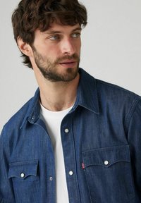 Camisa de denim azul con cuello puntiagudo, botones a presión y dos bolsillos frontales. Presenta un tejido texturizado y un sutil acento de logo rojo.