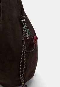 Brązowa zamszowa torba o miękkiej fakturze, wyposażona w metalowy klips oraz ozdobne sznurki w kolorach zielonym i czerwonym, a także ciemną skórzaną metkę.