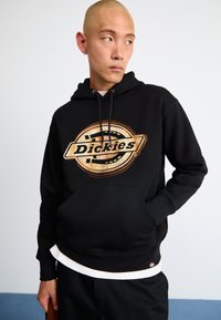 Μαύρη μπλούζα με κουκούλα που διαθέτει μια μεγάλη κυκλική γραφική παράσταση με την ένδειξη "Dickies" σε έντονη γραφή. Έχει εμπρόσθια τσέπη και ριμπ τελειώματα, η οποία είναι επικαλυμμένη από λευκό στρίφωμα.