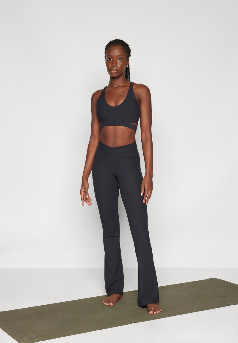 Sostén deportivo negro con un diseño recortado, combinado con leggings negros de campana de cintura alta. La modelo está de pie sobre una esterilla de yoga verde.