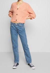 Persikorang stickad cardigan med ribbat mönster och stora knappar, i kombination med ljusblå raka jeans och svarta canvas sneakers.