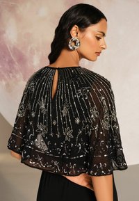 Juodas permatomas apsiaustas su sudėtinga sidabrine siuvinėjimu ir karoliukų dekoracijomis. Detalė su raktų skylute gale ir raukšlėta kraštinė. Sklandus, subtilus tekstūros visame kūne.