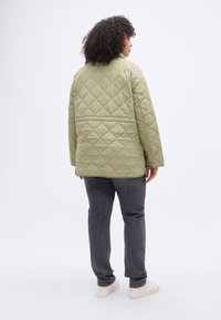 Καπιτονέ, απαλό χακί πράσινο μπουφάν με χαλαρή εφαρμογή, με όρθιο γιακά και άνοιγμα στην πλάτη, συνδυασμένο με σκούρο γκρι παντελόνια και λευκά αθλητικά παπούτσια.